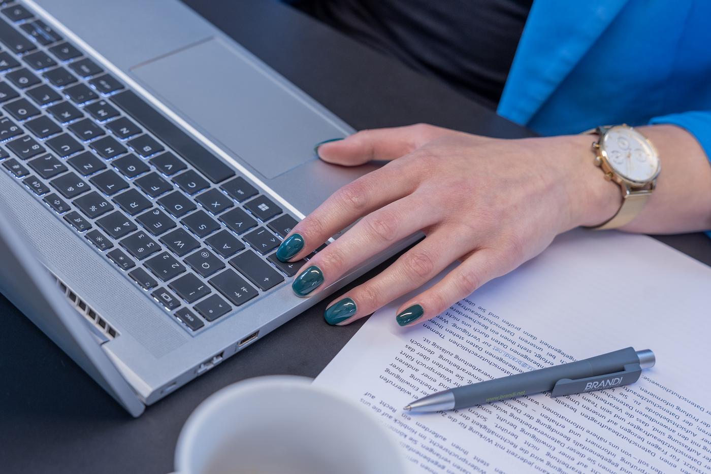 Eine Hand liegt halb auf einem Laptop. Daneben liegt eine Blatt und ein Kugelschreiben.